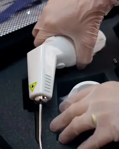 Mão de luva segurando o Alltherapy, com o aviso de laser no cabo e a ponta metálica (quadro de vídeo do Estevão)